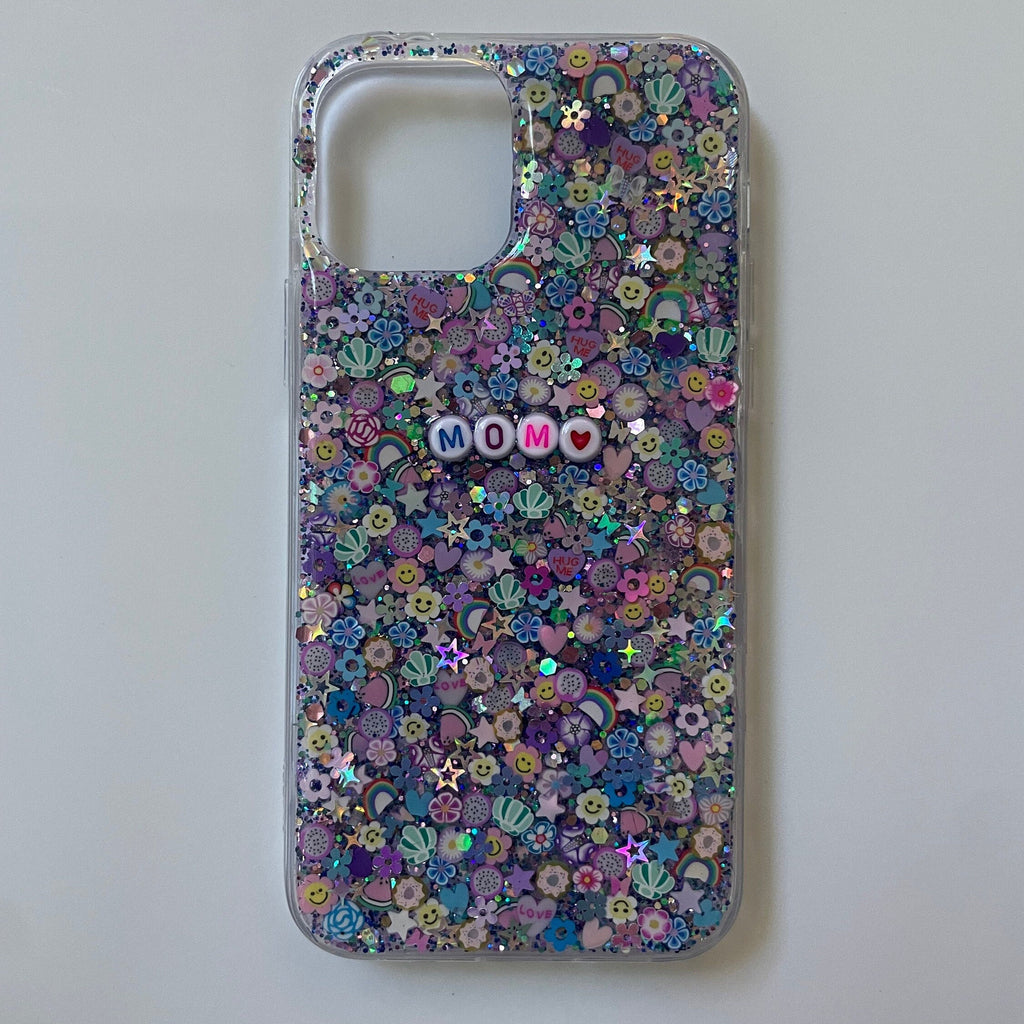 
A clear smartphone case featuring a maximalist, multi-colored design on a purple and teal glitter base. The back is densely packed with 3D charms, including strawberries, rainbow arches, flowers, and holographic stars. White circular beads with multicolored letters are centered on the case, spelling out the name "MOM." The case is shown against a white background with a large rectangular camera cutout at the top.