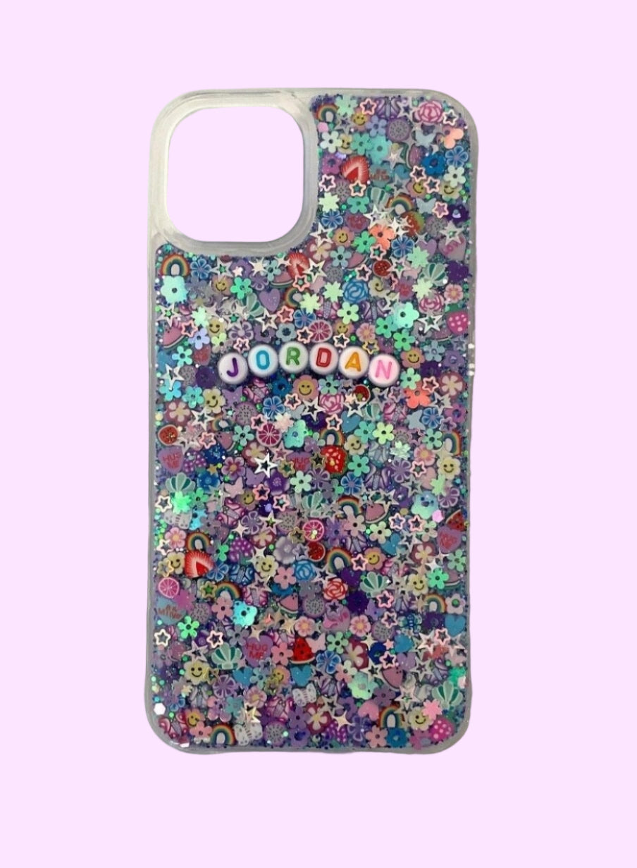 A clear smartphone case featuring a maximalist, multi-colored design on a purple and teal glitter base. The back is densely packed with 3D charms, including strawberries, rainbow arches, flowers, and holographic stars. White circular beads with multicolored letters are centered on the case, spelling out the name "JORDAN." The case is shown against a soft lavender background with a large rectangular camera cutout at the top.