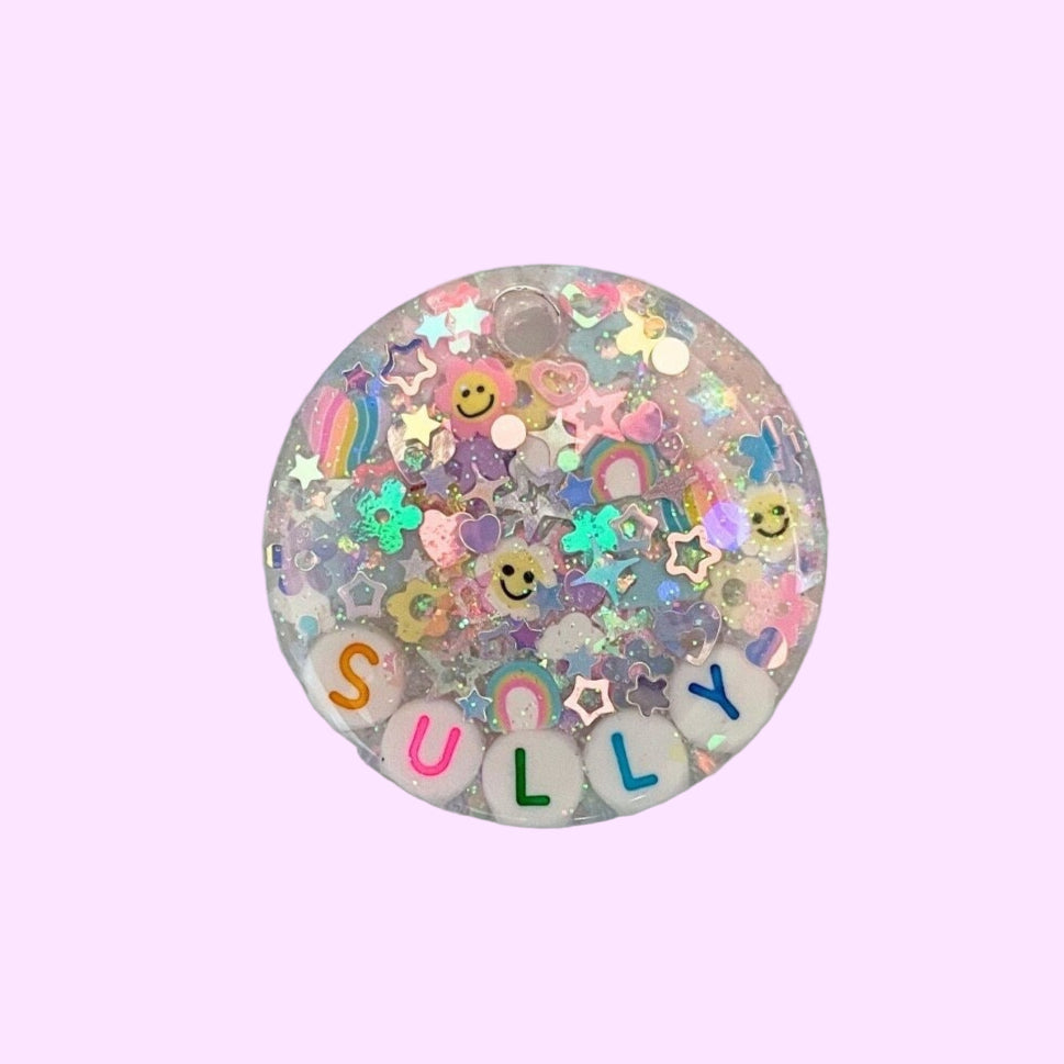 Round decorative item with iridescent glitter and letters spelling 'SULLY' on a holographic background